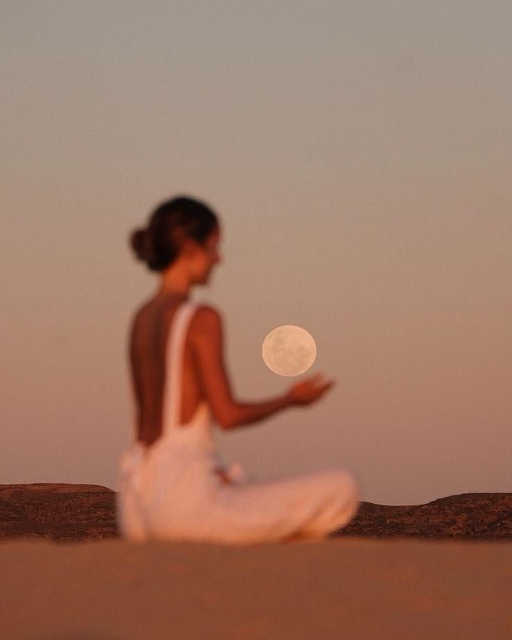 A woman in a white dress sitting in the desert at dusk, holding her hand beneath the full moon