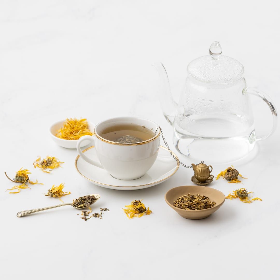 A cup of herbal tea beside a glass teapot, with dried calendula flowers and loose-leaf tea