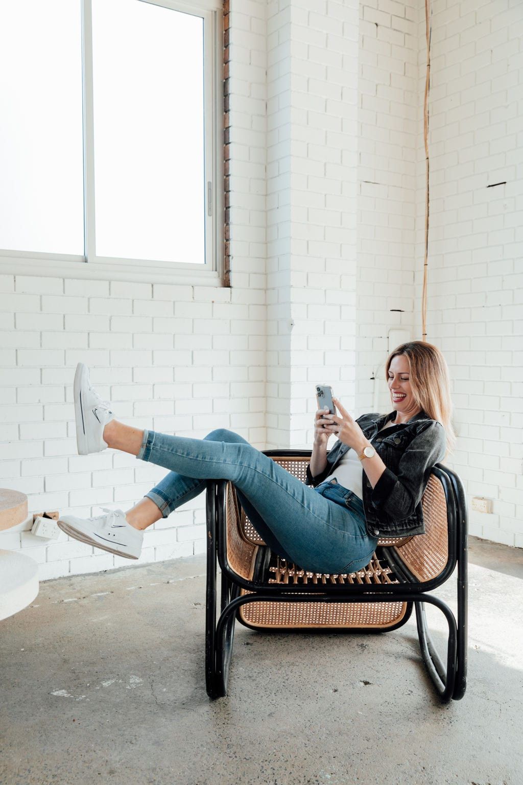 Dee Zibara laughing at her phone, sitting in a cane chair with her feet up